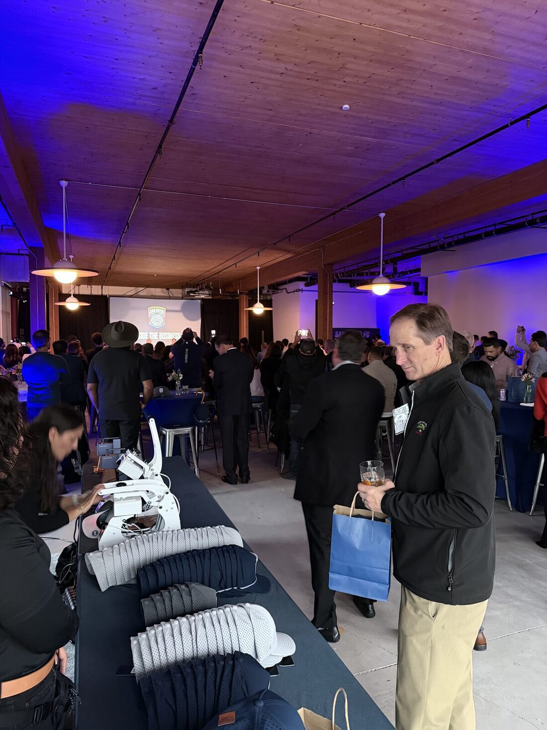 Guests at a San Francisco evening event near a merch table of folded caps under blue ceiling uplighting