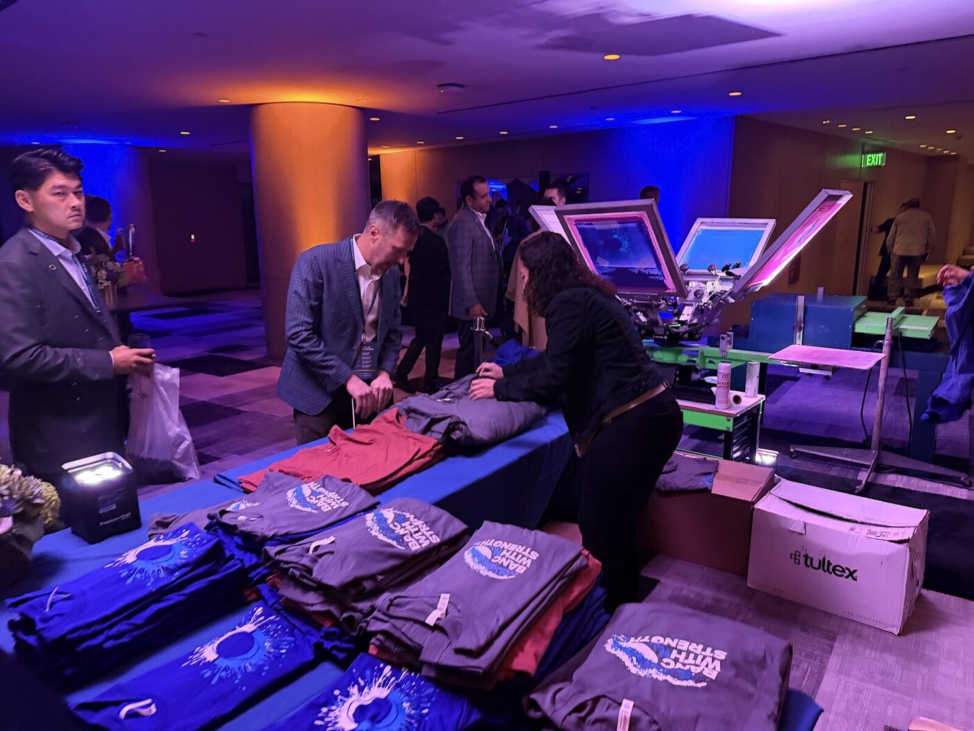 Live screen-printing press at a San Francisco corporate event as guests browse folded shirts under blue and amber lighting